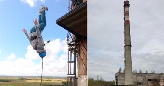 A person wearing a helmet and harness jumps off the side of a tall industrial building, upside down in mid-air; beside is a tall, old smokestack rising above an abandoned structure.