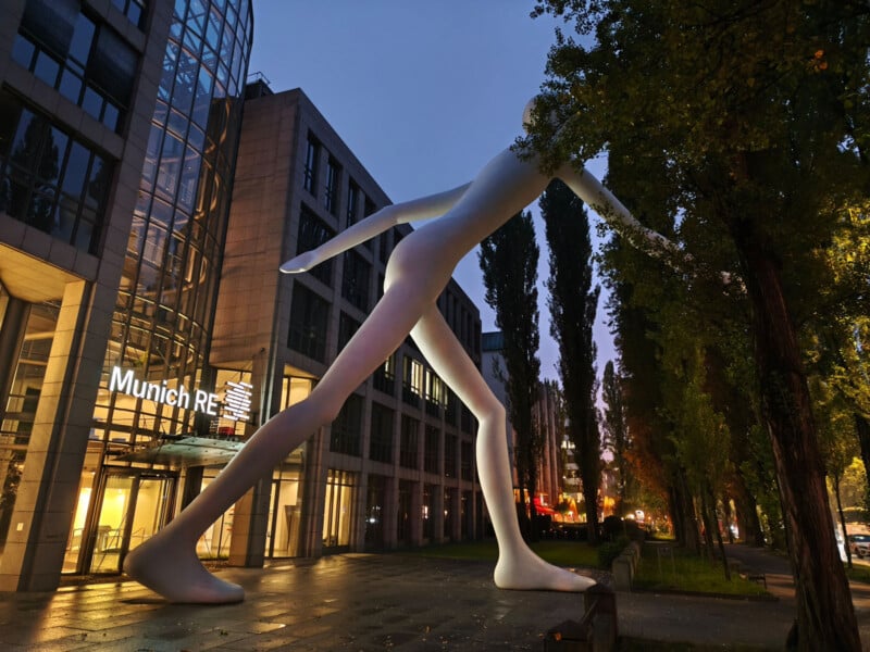A large, modern sculpture of a walking figure stands outside a glass-fronted office building with the sign "Munich Re" in an urban area, surrounded by tall trees and illuminated at dusk.