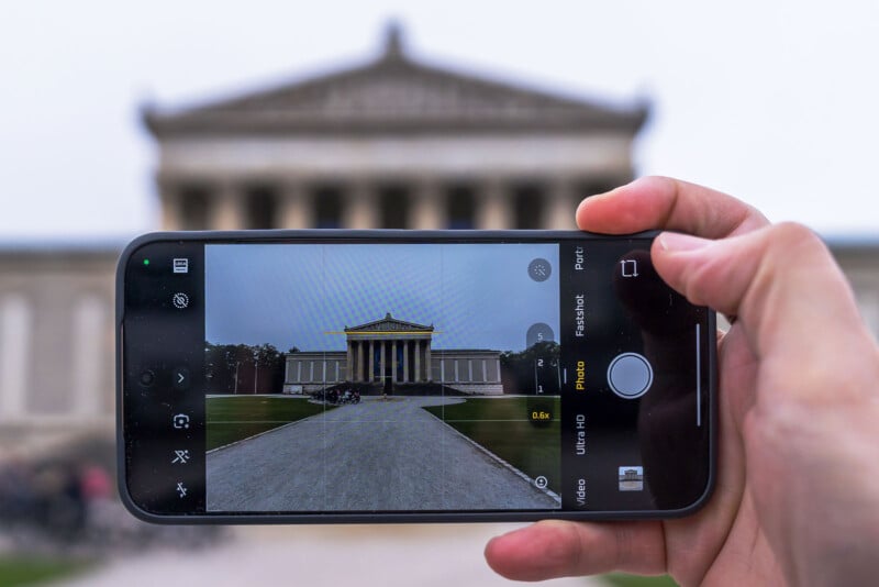 A person holds a smartphone in landscape mode, photographing a classical-style building with columns and a triangular pediment. The same building is visible both on the phone screen and in the background.