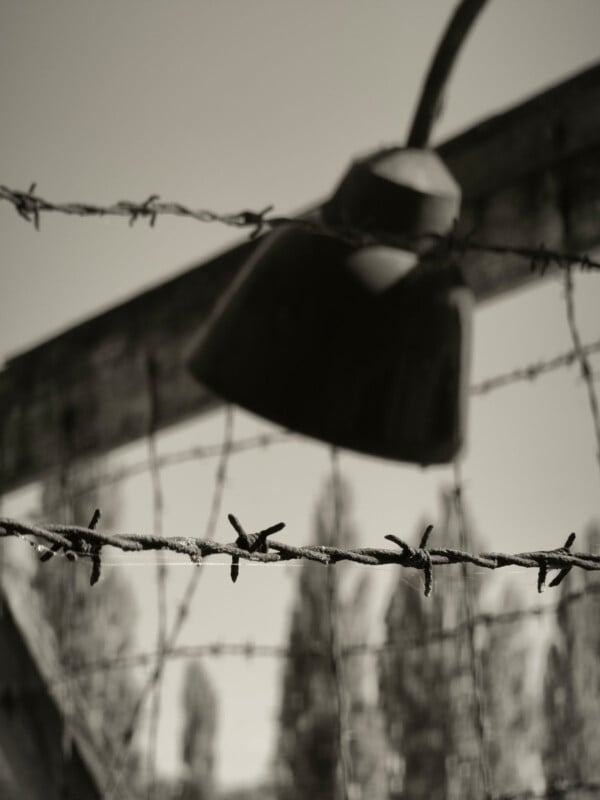 Close-up of barbed wire in focus with an old, out-of-focus lamp and wooden structure in the background; the scene is in black and white, evoking a somber, historical atmosphere.
