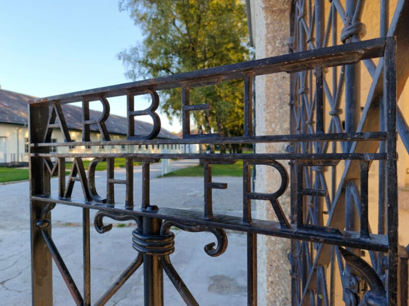 A close-up of an iron gate with the inscription "Arbeit macht frei" at the entrance of a concentration camp, with trees and buildings visible in the background.