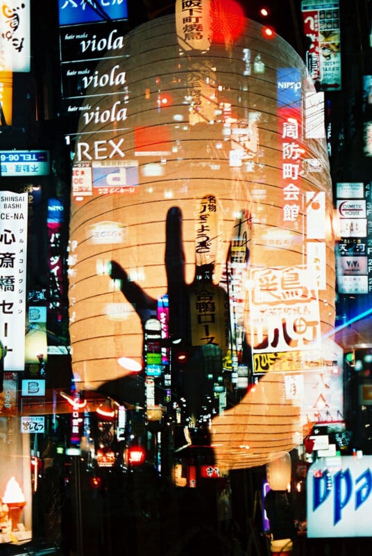 A hand silhouette is superimposed over a vibrant city scene filled with colorful neon signs and Japanese characters, creating a layered, visually striking effect.