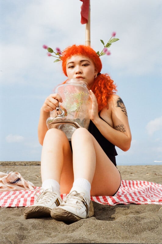 A person with bright orange hair, floral hair accessories, and visible tattoos sits on a beach towel, holding a large glass jug. They wear a black outfit and white sneakers, with sand and sea in the background.
