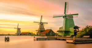 Three traditional Dutch windmills stand by the water at sunset, with colorful clouds in the sky. Wooden docks and small houses are visible near the windmills, surrounded by calm water and lush greenery.