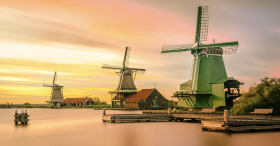 Three traditional Dutch windmills stand by the water at sunset, with colorful clouds in the sky. Wooden docks and small houses are visible near the windmills, surrounded by calm water and lush greenery.