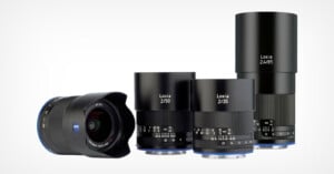 Four Zeiss Loxia camera lenses are displayed standing side by side on a white background, showing their model numbers: 2/21, 2/50, 2/35, and 2.4/85. The lens on the left is angled forward.