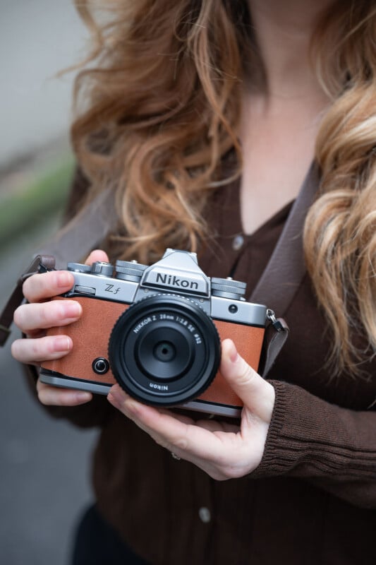 A woman with long, wavy blonde hair holds a vintage-style Nikon Z fc camera with both hands. She is wearing a brown shirt, and her face is partially out of frame.