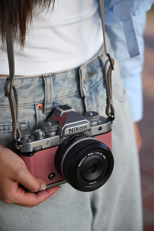 A person in blue jeans and a white top holds a pink Nikon Z f camera with a lens, supported by a strap around their neck. The camera rests near their waist.