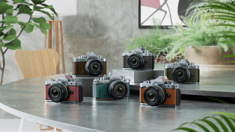 Six Nikon cameras in different colors are displayed on a round table with indoor plants, a wooden chair, and abstract art in the background, creating a stylish and modern scene.