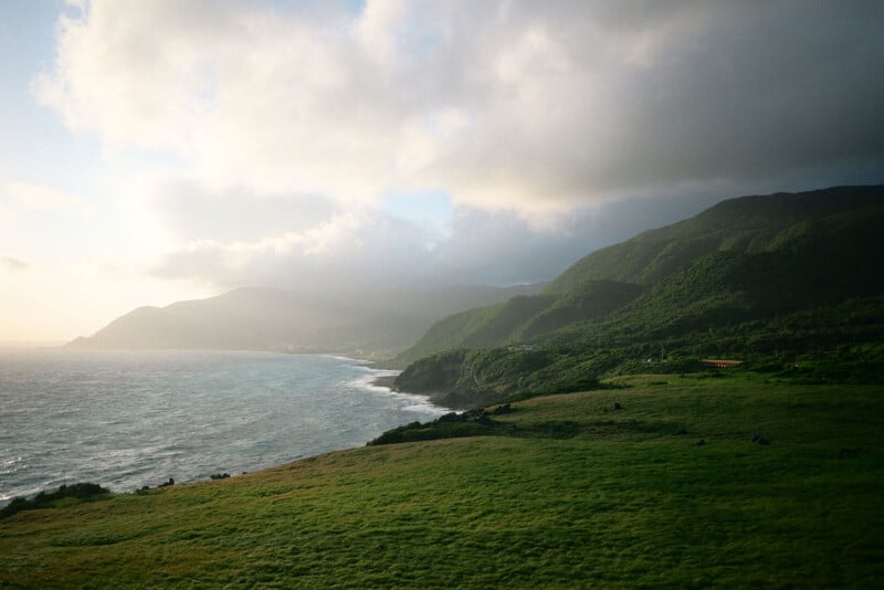 A coastal landscape with green hills meeting the ocean, waves crashing against the shore, and sunlight breaking through partly cloudy skies.