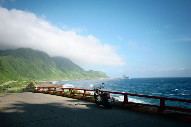 A scooter is parked by a red railing along a coastal road, overlooking the blue ocean and green mountains under a partly cloudy sky.