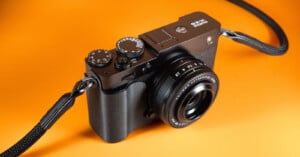 A black digital camera with a lens, control dials, and a strap, is positioned on an orange background.