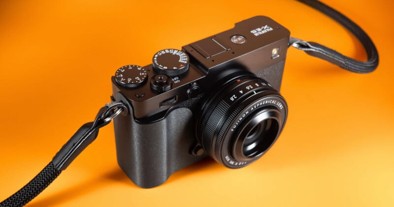 Una cámara digital negra con lente, dial de control y cinturón se encuentra sobre un fondo naranja.