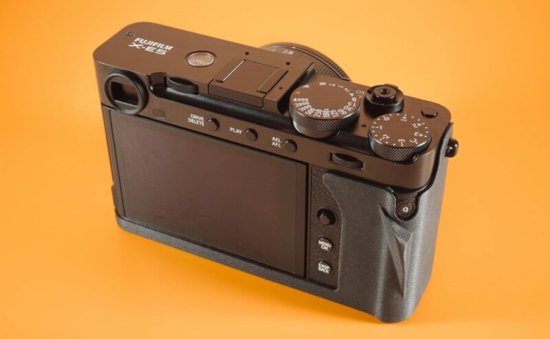Desde la parte posterior del fondo naranja, se muestra la cámara digital Fuji X-E3, que muestra su pantalla LCD, dial de control, botones y agarres texturizados.