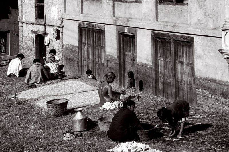 People are sitting and working outside a building with several closed doors; some are washing or preparing food, while others are socializing or working near a basket and metal containers on the ground.