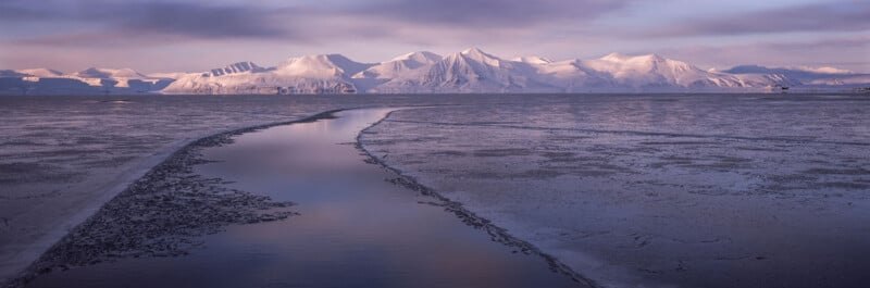 A narrow channel of water winds through an icy, frozen landscape toward snow-covered mountains under a pastel-colored sky at sunset or sunrise.