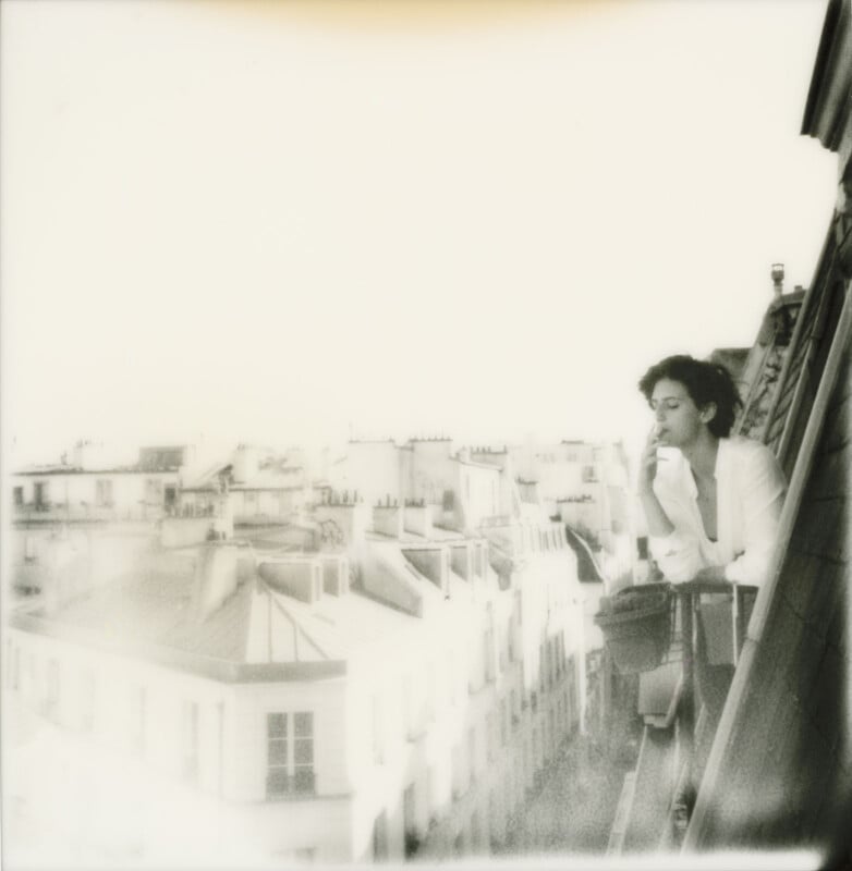 A person in a white shirt leans on a balcony railing, looking thoughtful while overlooking rooftops and buildings in a softly blurred, sunlit cityscape.