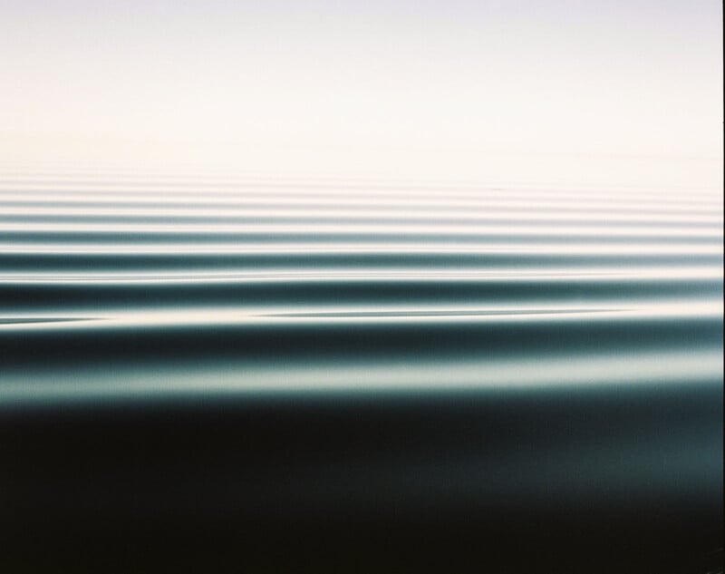 Calm, dark blue water with gentle, evenly spaced ripples extends to the horizon, blending into a pale, hazy sky. The overall scene is minimalistic and tranquil.