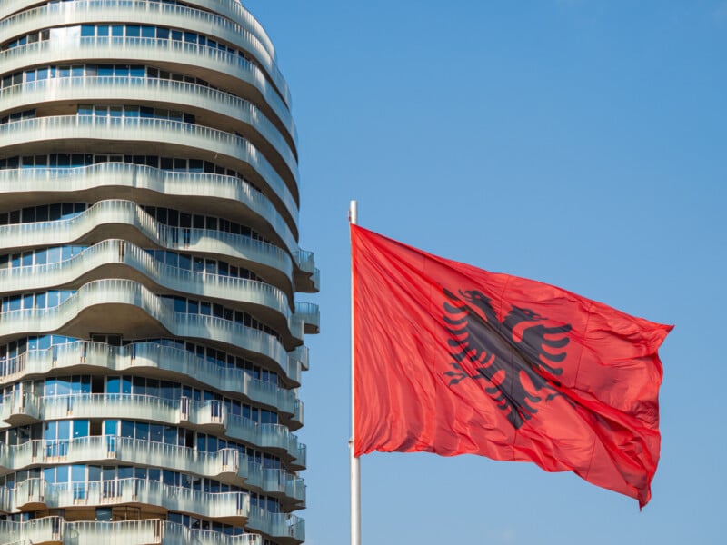 A red Albanian flag with a black double-headed eagle waves in front of a modern, curved glass building under a clear blue sky.