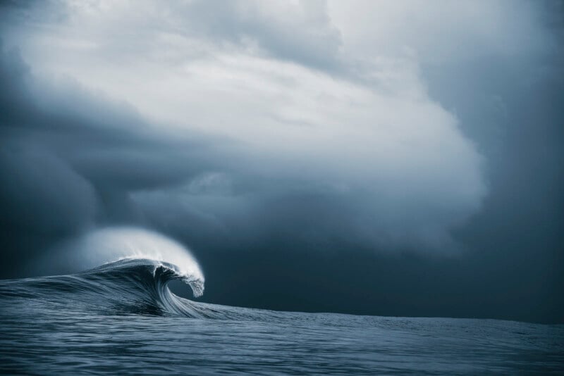 A large ocean wave curls dramatically under a moody, overcast sky, with dark clouds looming in the background and soft light highlighting the crest of the wave.