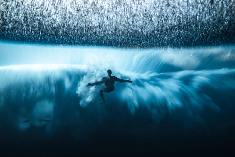 A surfer is underwater, surrounded by swirling blue waves and bubbles after wiping out, with light filtering through the water above.