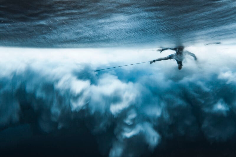 A surfer is underwater, partially visible through the surface as powerful, foamy waves churn beneath them, creating a dramatic and blurred blue and white scene.