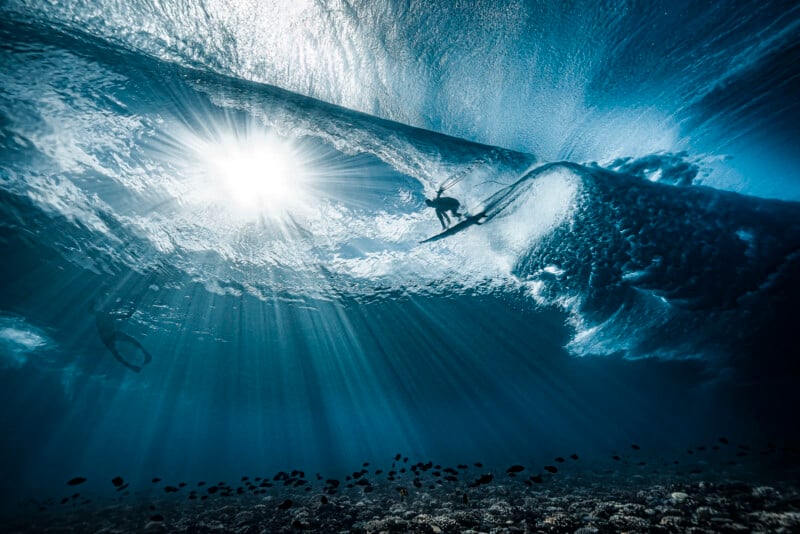 Underwater view of a surfer riding a wave with sunlight streaming through the water. The wave curls above, creating dramatic patterns, and another person is visible swimming nearby. The ocean floor is visible below.