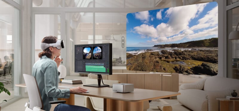 A person wearing a VR headset sits at a desk editing video on a computer with two monitors. A large virtual display shows a scenic rocky coastline with blue sky and clouds inside a modern office.