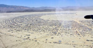 Aerial view of a large, semicircular city of tents, vehicles, and temporary structures set up on a vast desert plain, surrounded by mountains in the distance under a clear blue sky.