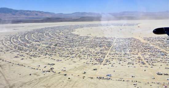 Aerial view of a large, semicircular city of tents, vehicles, and temporary structures set up on a vast desert plain, surrounded by mountains in the distance under a clear blue sky.
