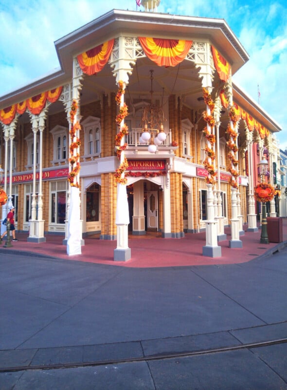 A large, ornate building decorated with orange and yellow bunting and wreaths for fall, featuring columns, arches, and a sign reading "Emporium" above the entrance. The sky is blue with some clouds.