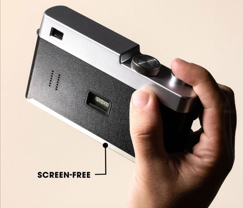 A hand holds a compact, retro-style camera with a black and silver design. A label points to the back, highlighting that the camera is "screen-free" with no visible display.