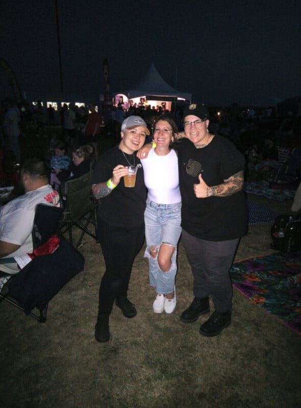 Three people smile and pose together at an outdoor nighttime event. Two wear black shirts and hats, while the person in the middle wears a white top and ripped jeans. A crowd and tented stage with lights are visible in the background.
