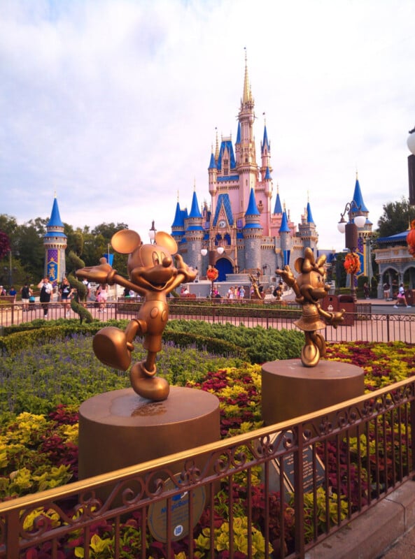 Golden statues of Mickey and Minnie Mouse stand in front of a flower bed, with the Cinderella Castle and its blue turrets rising in the background at a Disney theme park, under a partly cloudy sky.