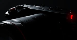 Close-up of a black camera body and lens with a textured grip, illuminated by low light, highlighting a red button and a red indicator light. The scene is mostly shadowed, emphasizing the camera’s sleek design.
