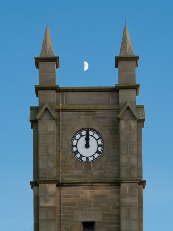 Una torre de reloj de piedra con una aguja muestra la hora a las 6:00, contra un cielo azul claro, con la media luna directamente encima de la torre.