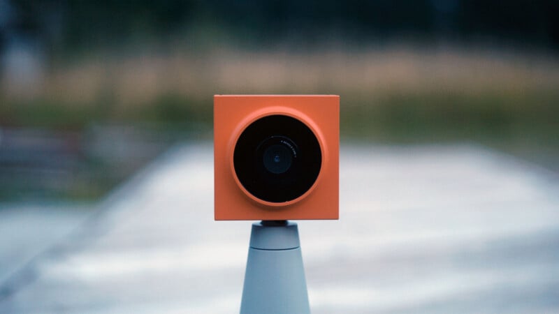 An orange, square-shaped camera sits atop a gray stand, facing directly forward, with a blurred outdoor background of grass and trees.