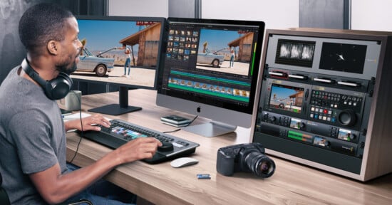 A man sits at a desk editing a video on a large computer; he wears headphones and uses a keyboard with colored keys. Nearby are a camera, an SD card, control panels, and two monitors displaying video footage.