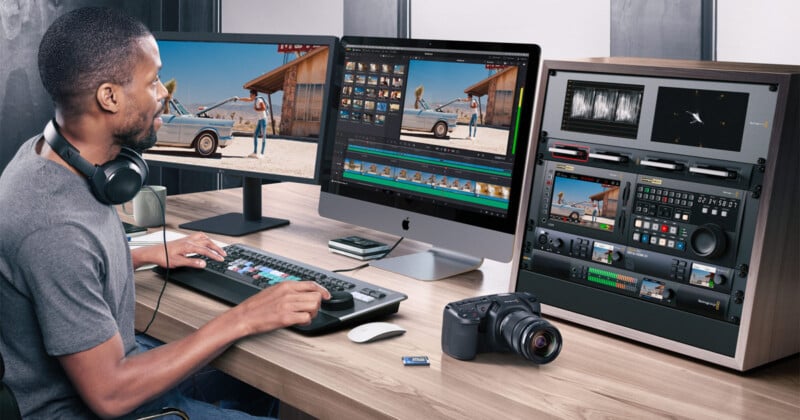 Un hombre sentado en un escritorio editando video en una computadora grande. Lleva auriculares y usa un teclado con una tecla de color. Cerca están la cámara, una tarjeta SD, un panel de control y dos monitores que muestran la lente de video.