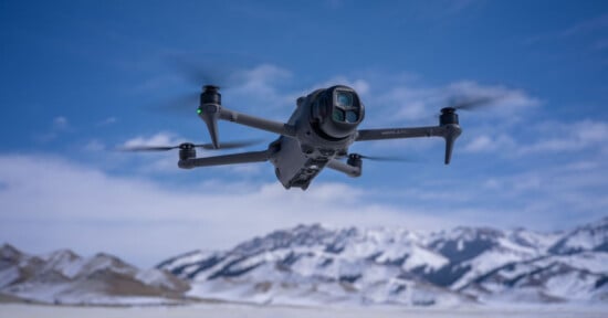 A black quadcopter drone flying in a clear blue sky with snow-covered mountains and a snowy landscape in the background.