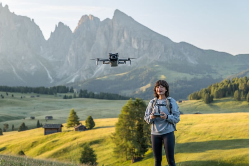A woman operates a drone with a remote control in a scenic, sunlit valley with rolling green hills, scattered cabins, and tall mountains in the background.