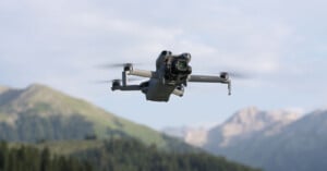 A camera drone flies in the air with blurred mountains and trees in the background under a partly cloudy sky.