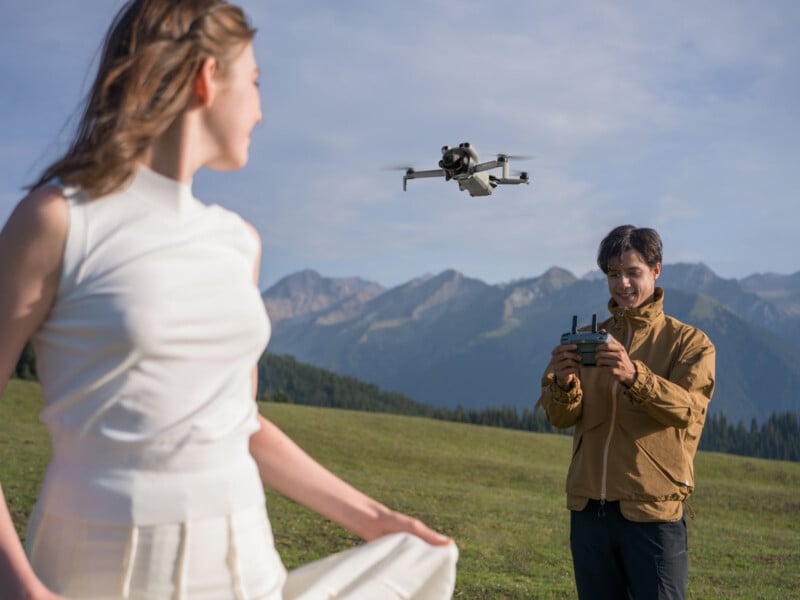 A man in a brown jacket flies a drone with a remote control in a grassy field with mountains in the background, while a woman in white stands in the foreground, slightly out of focus.