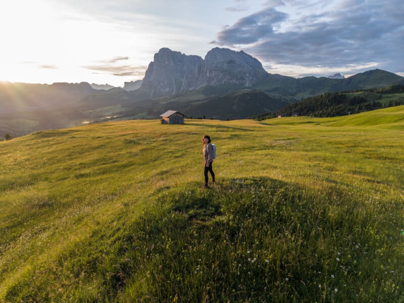A person stands on a grassy field at sunset with mountains and a small cabin in the background under a partly cloudy sky.