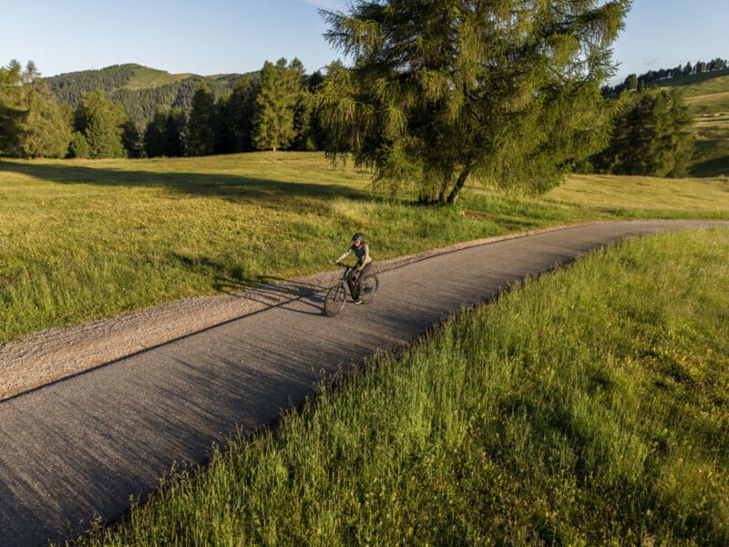 A person rides a bicycle on a paved path through a sunlit grassy field with trees and rolling hills in the background. The scene is peaceful and bathed in warm, late afternoon light.