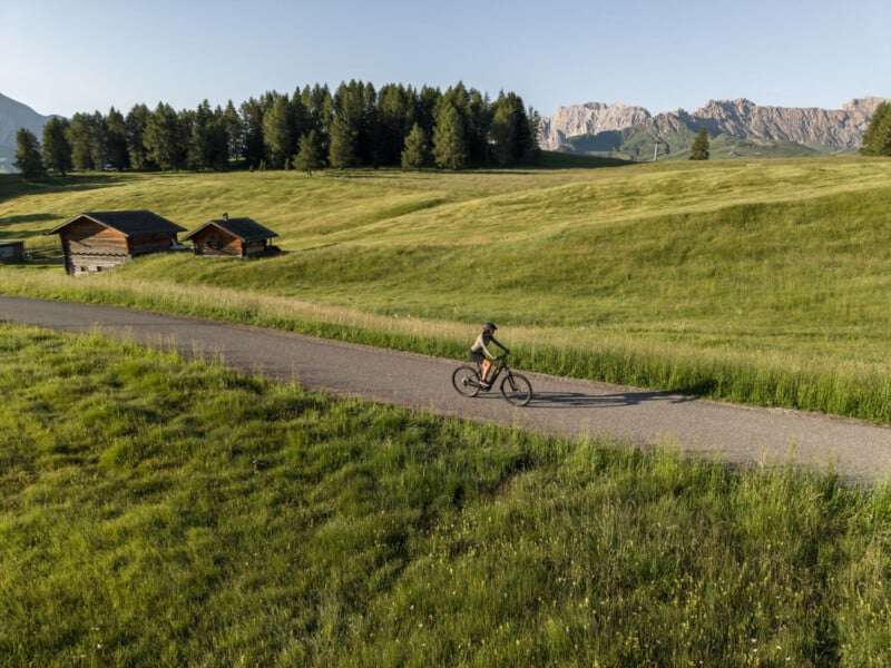 A cyclist rides along a paved path through a green meadow with scattered wooden cabins, pine trees, and distant mountains under a clear sky.