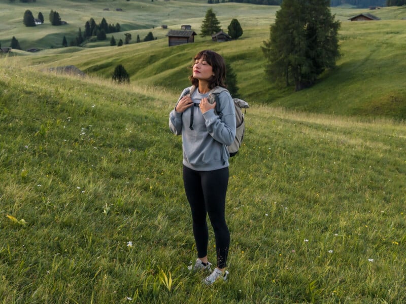 A woman wearing a gray hoodie and black leggings stands on a grassy hillside with her eyes closed, enjoying the outdoors. She has a backpack and is surrounded by green fields, trees, and a few wooden cabins in the distance.