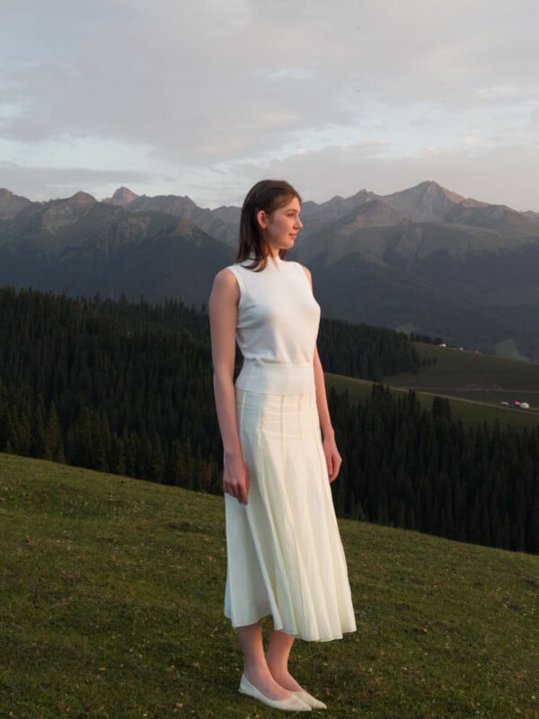 A woman in a sleeveless white top and long white skirt stands on grassy hillside, looking to the side, with pine forests and mountain peaks in the background under a cloudy sky.