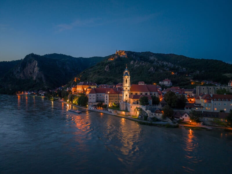 A riverside village at dusk with an illuminated church tower, red-roofed buildings, and hills in the background, reflecting softly on the calm water.