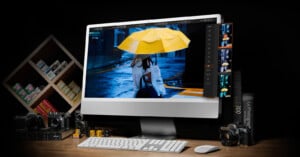 A desktop computer displays a photo of a person holding a yellow umbrella in the rain. Surrounding the monitor are film cameras, photo film rolls, photography books, and a shelf with film boxes, all set on a wooden desk.
